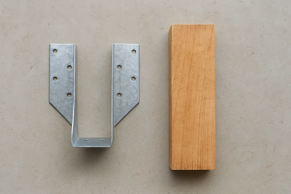 Two plain galvanised steel joist hangers on a bare concrete slab beside a short length of timber — patio cover, Longview. Illustrative example.